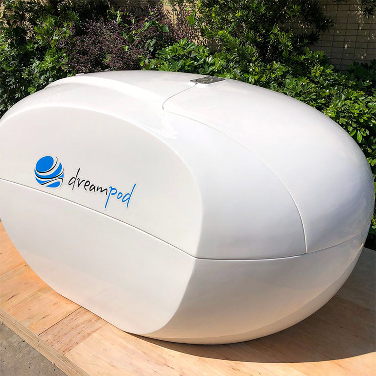 Dreampod Home Float Plus, a sleek white flotation pod, displayed outdoors with greenery in the background.