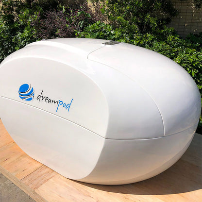 Dreampod Home Float Plus, a sleek white flotation pod, displayed outdoors with greenery in the background.