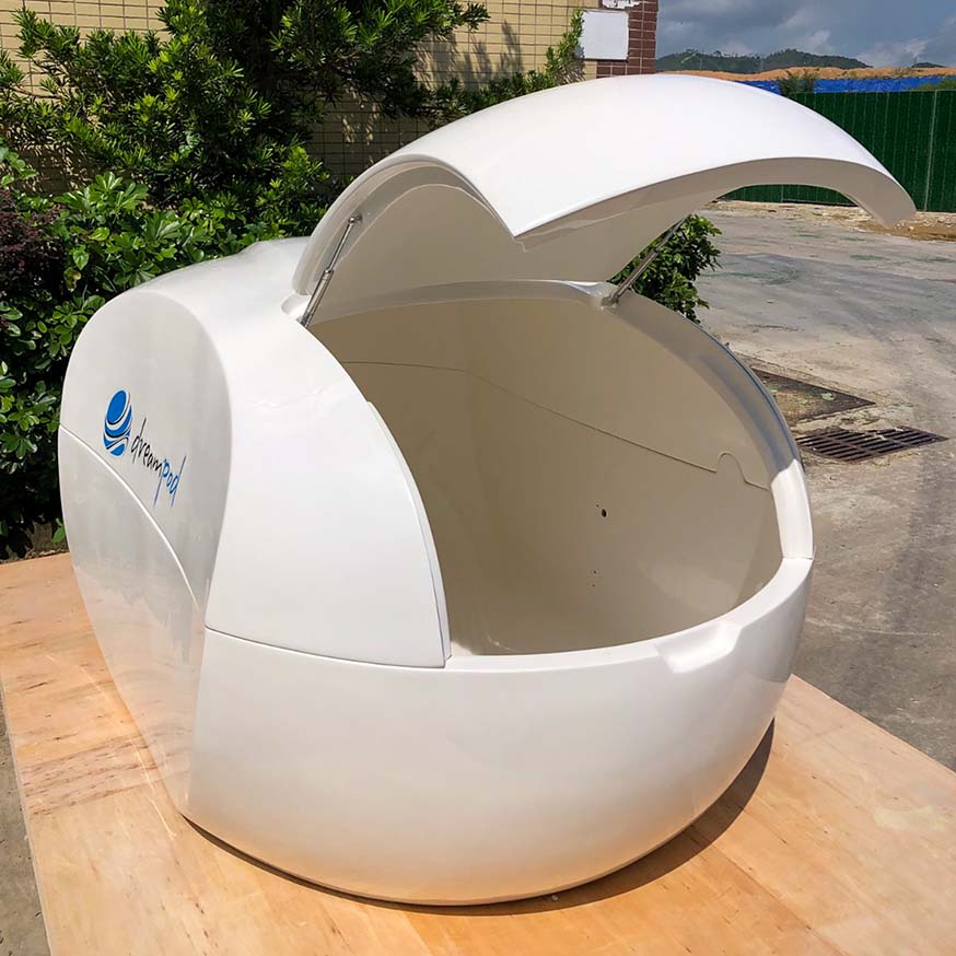 Dreampod Home Float Plus, a sleek white floatation pod with an open lid, displayed on a wooden surface surrounded by greenery.