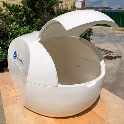 Dreampod Home Float Plus, a sleek white floatation pod with an open lid, displayed on a wooden surface surrounded by greenery.