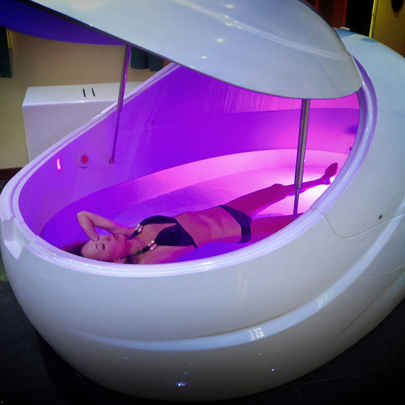 A person relaxing inside a Dreampod V2 Float Pod, illuminated with soft purple lighting.