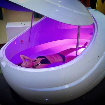 A person relaxing inside a Dreampod V2 Float Pod, illuminated with soft purple lighting.