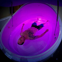 A person floating in a Dreampod V2 Float Pod filled with water, illuminated by soft purple lights.
