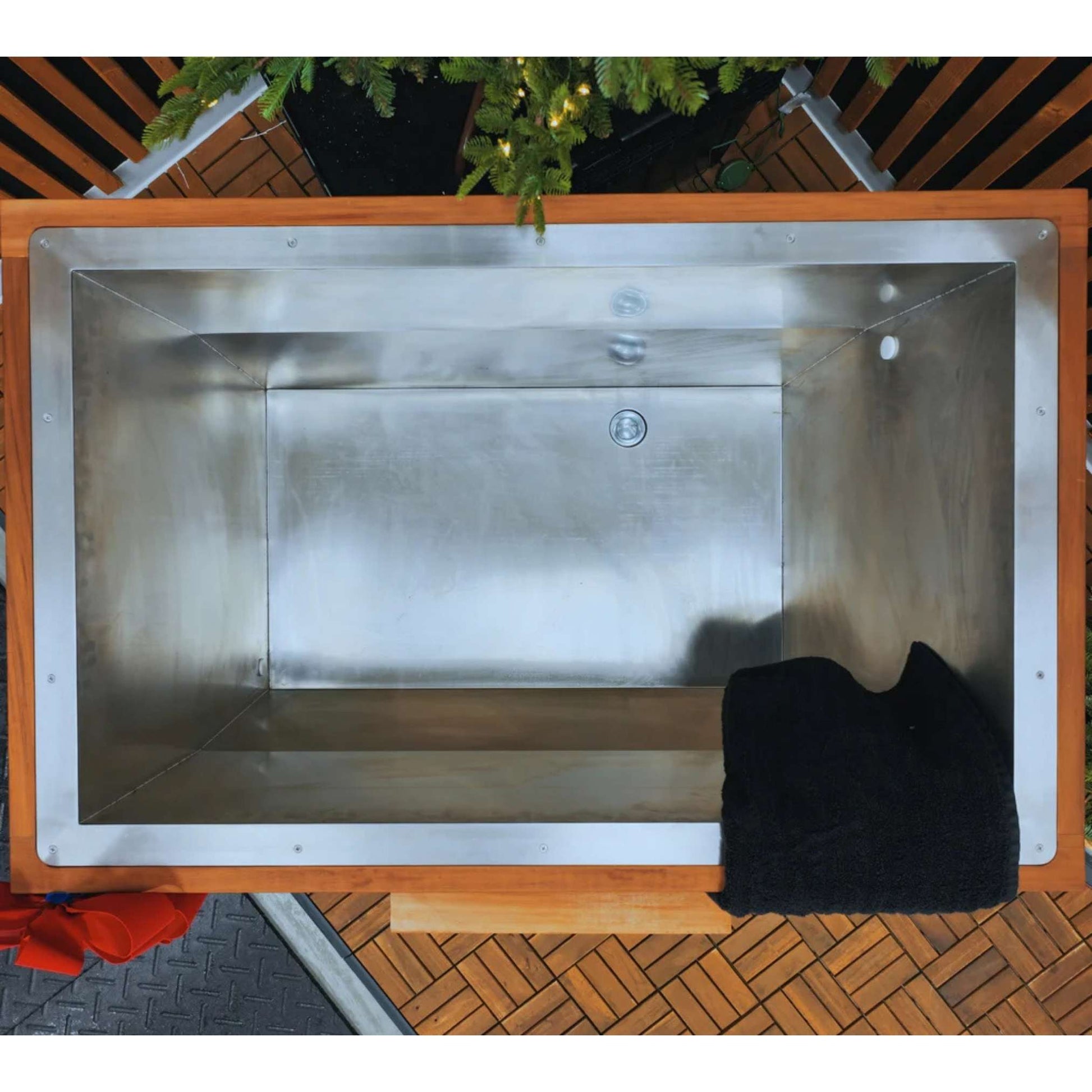 An overhead view of a stainless steel tub with a wooden frame, featuring a black towel resting on the edge. The background includes greenery and a wooden deck.