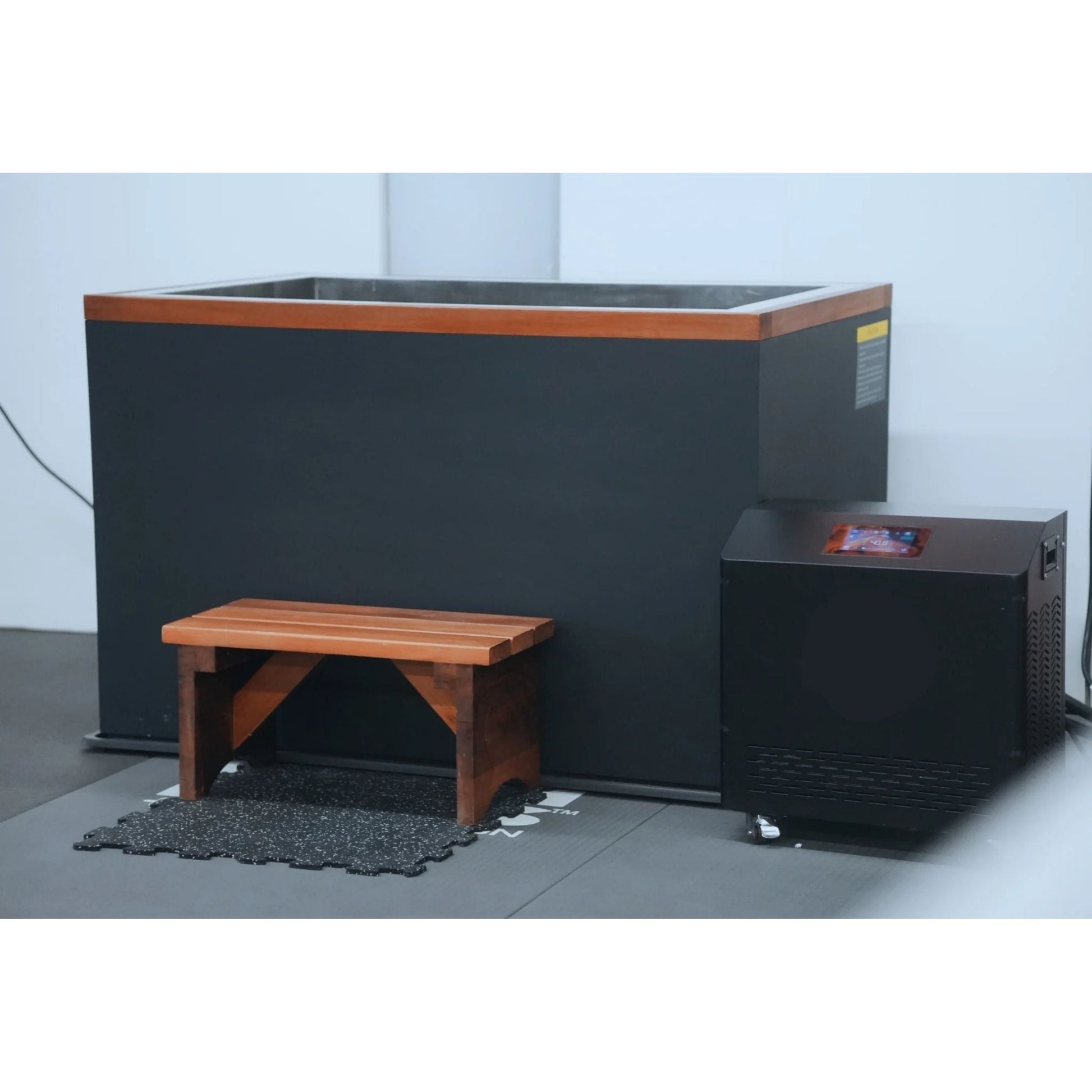 A modern cold plunge tub with a wooden top and a wooden bench in front, accompanied by a black cooling unit on the right, set against a minimalist indoor background.