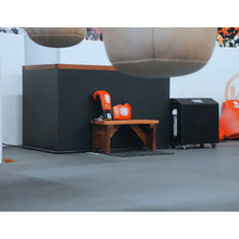 A training area featuring a black counter with a wooden bench in front. On the bench are two orange boxing gloves labeled EDDY and RAMIREZ. In the background, there's a black cabinet and soft flooring.