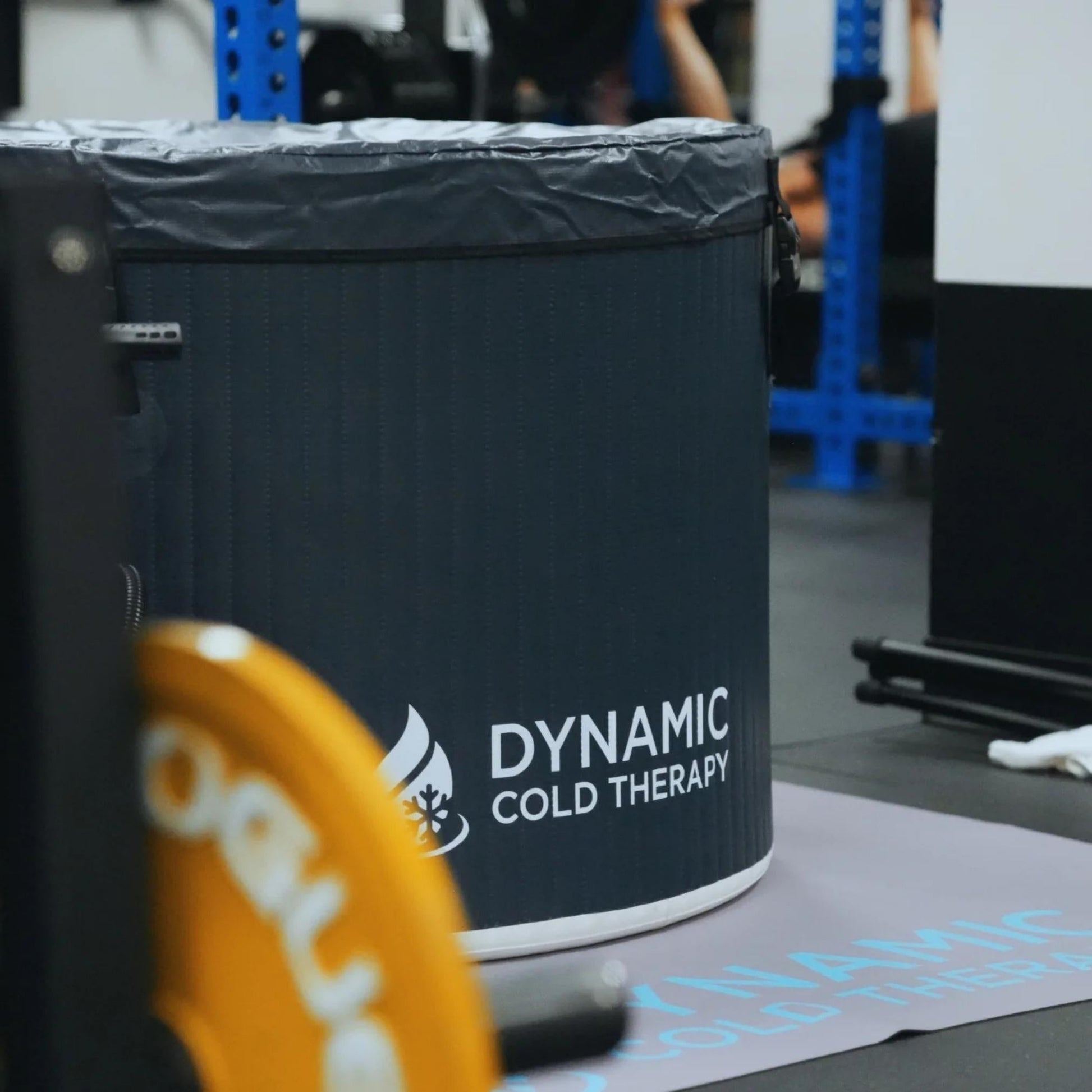 A close-up view of a Dynamic Cold Therapy tub in a gym setting, with a blurred weight plate in the foreground and workout equipment visible in the background.