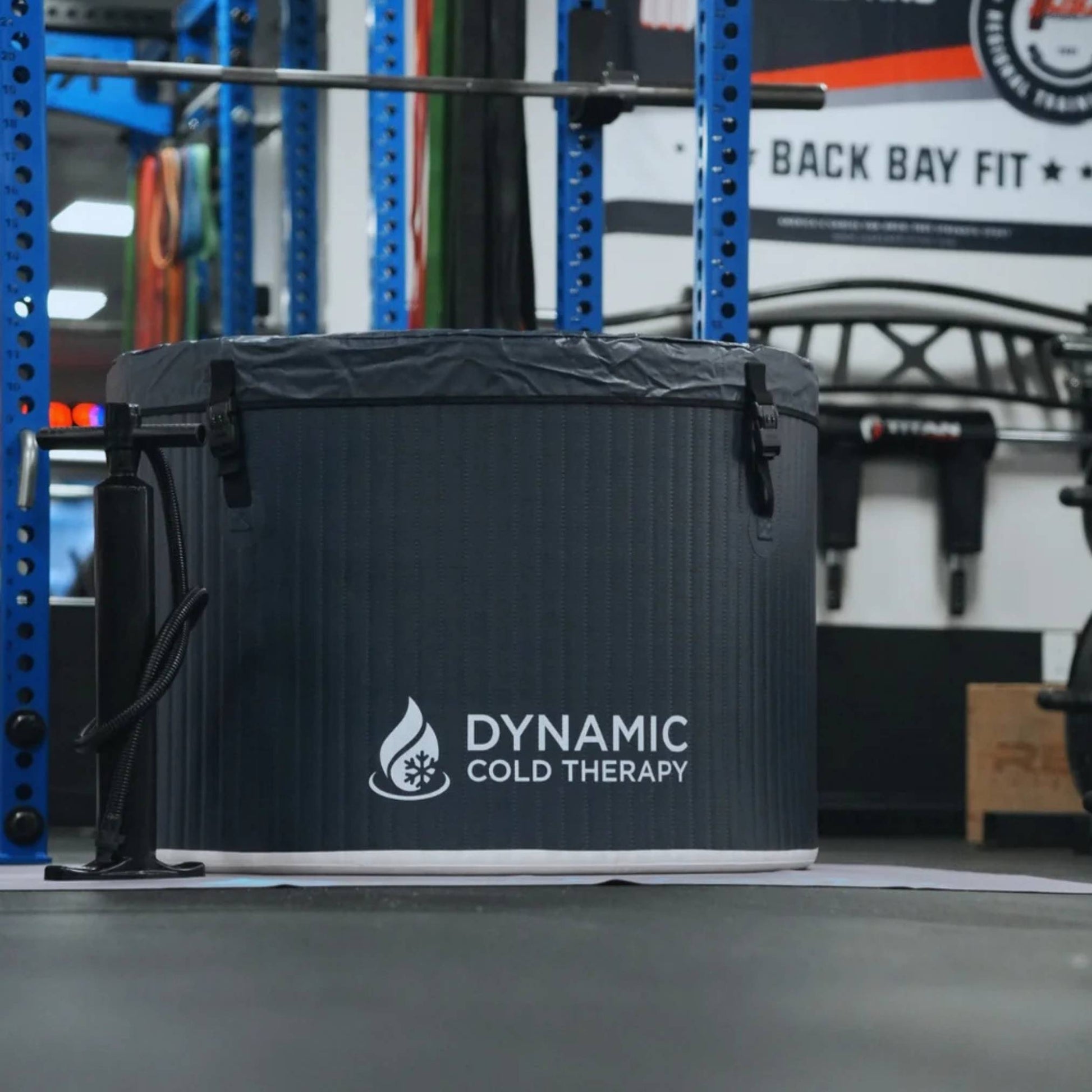 A Dynamic Cold Therapy portable ice bath with a black exterior, positioned in a gym environment featuring blue equipment and fitness accessories in the background.