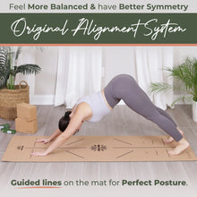 Woman in downward dog pose on a natural cork yoga mat with alignment guide lines printed across the surface, set in a bright room with potted plants and wooden props.