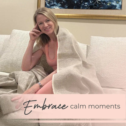 Woman sitting on a cream-colored sofa draped with a beige hemp-organic cotton yoga blanket, smiling at camera in a home setting.