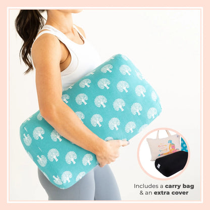 Woman holding a turquoise yoga bolster pillow with white tree pattern, wearing white sports bra and gray leggings. Includes carry bag and extra removable cover shown in circular inset.