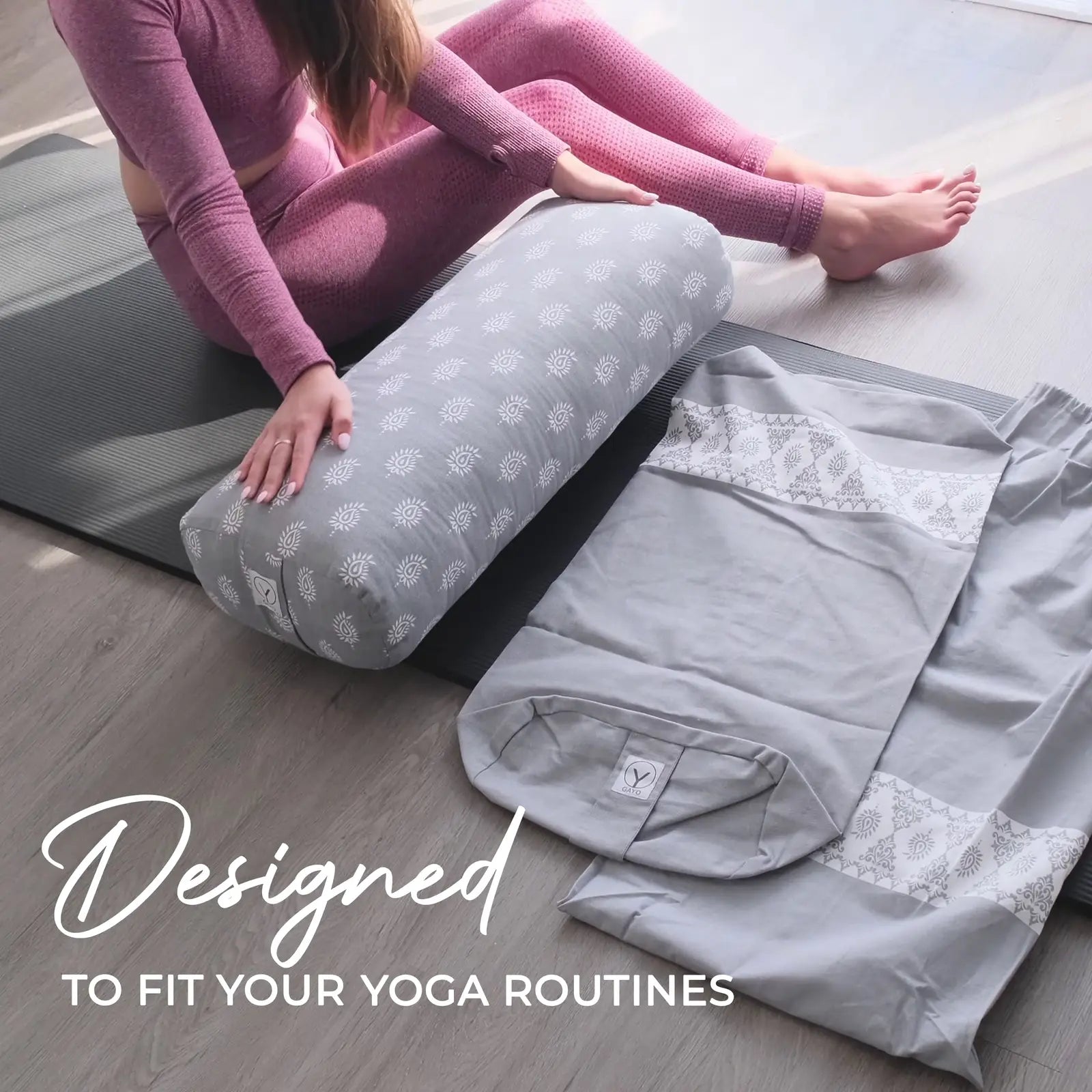 Woman in purple workout clothing resting her legs on a gray cylindrical yoga bolster with white sun pattern, alongside folded yoga accessories and a carry bag on a mat.