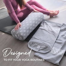 Woman in purple workout clothing resting her legs on a gray cylindrical yoga bolster with white sun pattern, alongside folded yoga accessories and a carry bag on a mat.