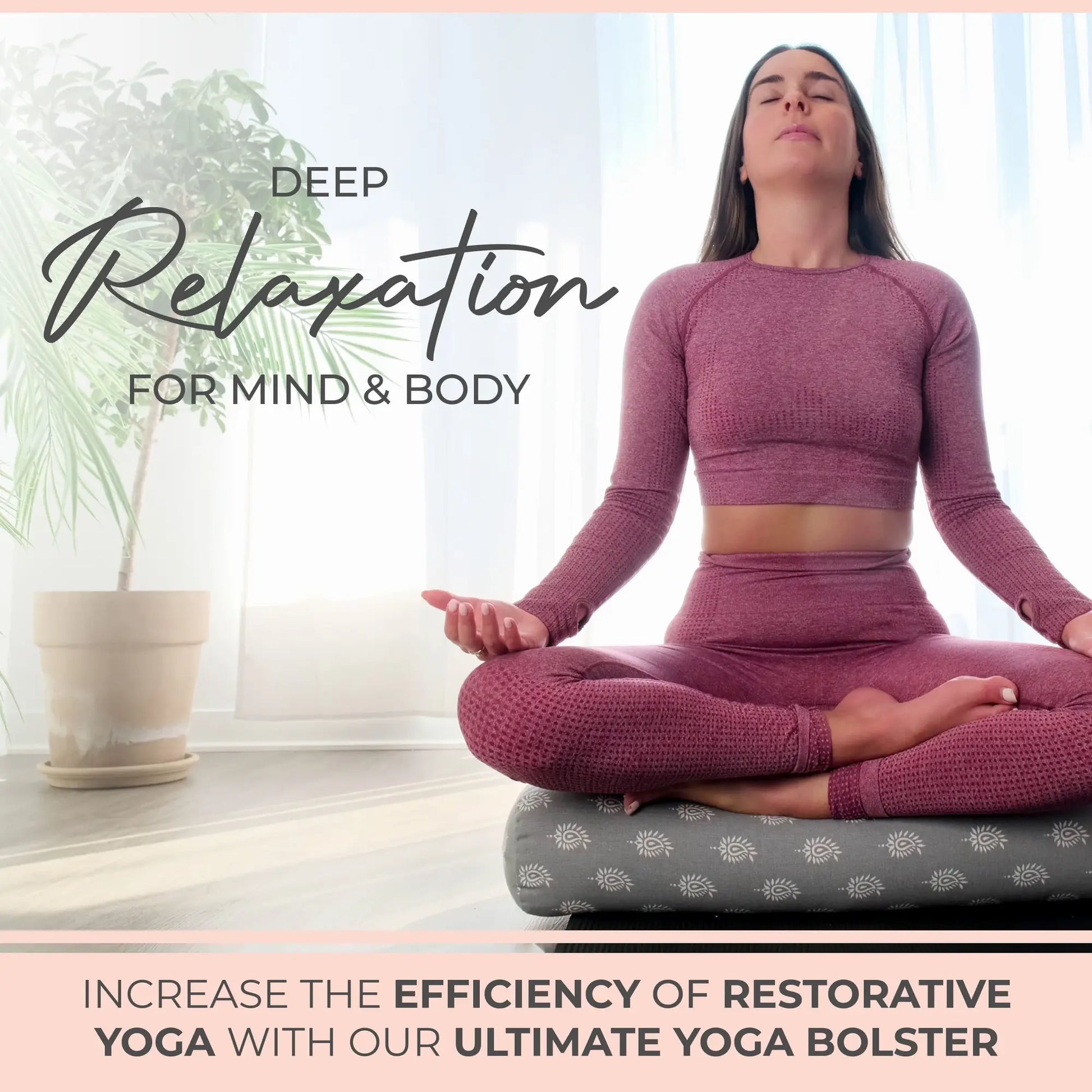 Woman in burgundy workout attire sitting in meditation pose on a gray yoga bolster with white pattern, next to a potted plant in bright natural light.