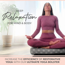 Woman in burgundy workout attire sitting in meditation pose on a gray yoga bolster with white pattern, next to a potted plant in bright natural light.