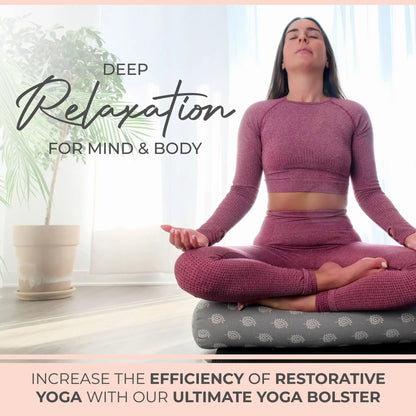 Woman in burgundy workout attire sitting in meditation pose on a gray yoga bolster with white pattern, next to a potted plant in bright natural light.