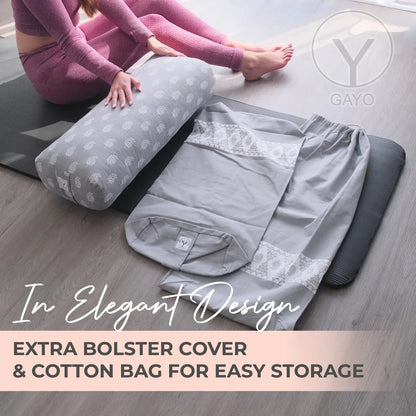 Woman in purple yoga attire sitting beside a gray cylindrical yoga bolster with white floral pattern and its matching cotton storage bag on a wooden floor.