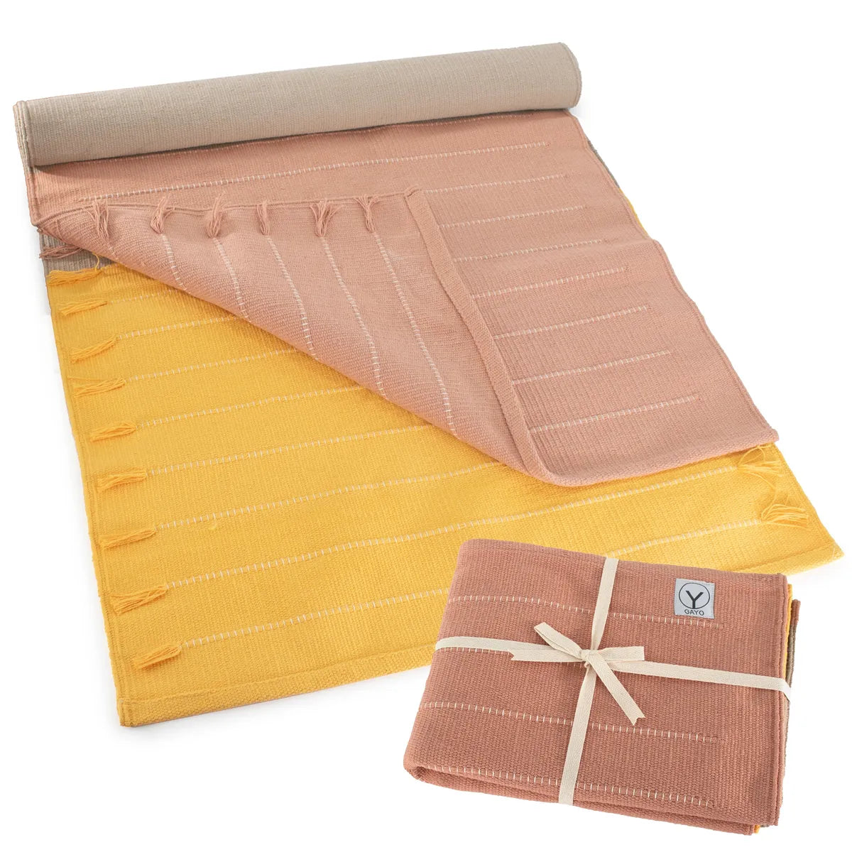 Rolled yoga mat in beige, pink, and yellow organic cotton with quilted texture and carrying strap; coordinating rolled towel in mauve pink tied with cream ribbon.