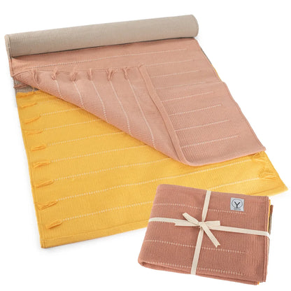 Rolled yoga mat in beige, pink, and yellow organic cotton with quilted texture and carrying strap; coordinating rolled towel in mauve pink tied with cream ribbon.