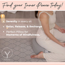 Woman in white clothing sitting in a meditative pose on a gray quilted buckwheat meditation cushion with a textured surface and brand logo visible.