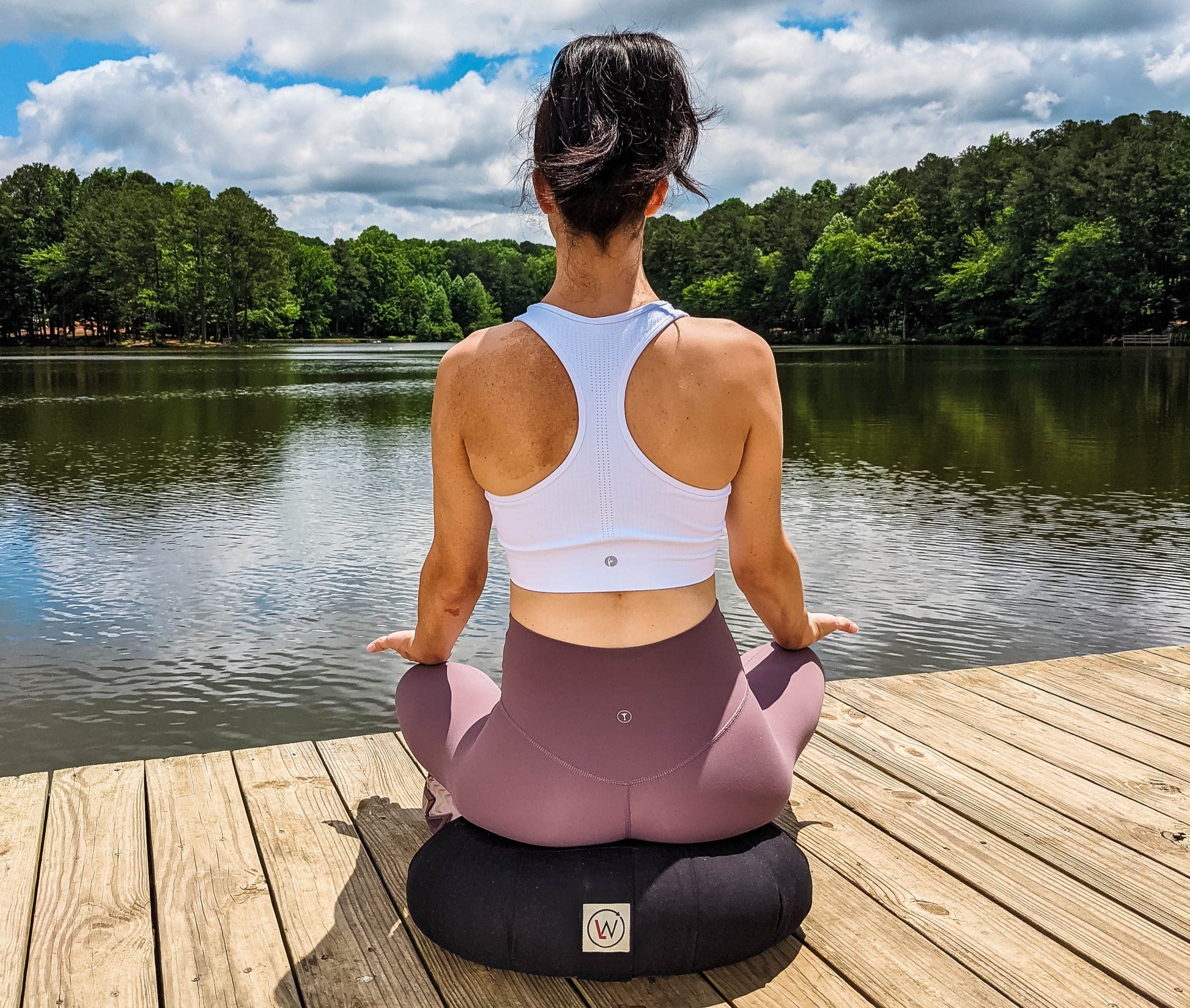 Woman meditating on a wooden dock by a lake; white sports top and mauve leggings.