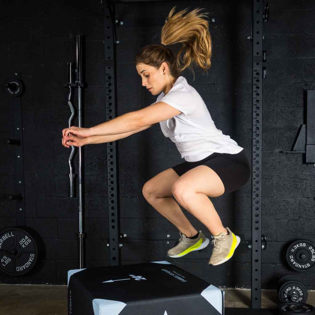 Woman doing a box jump in a gym, mid-air, wearing a white shirt and black shorts.