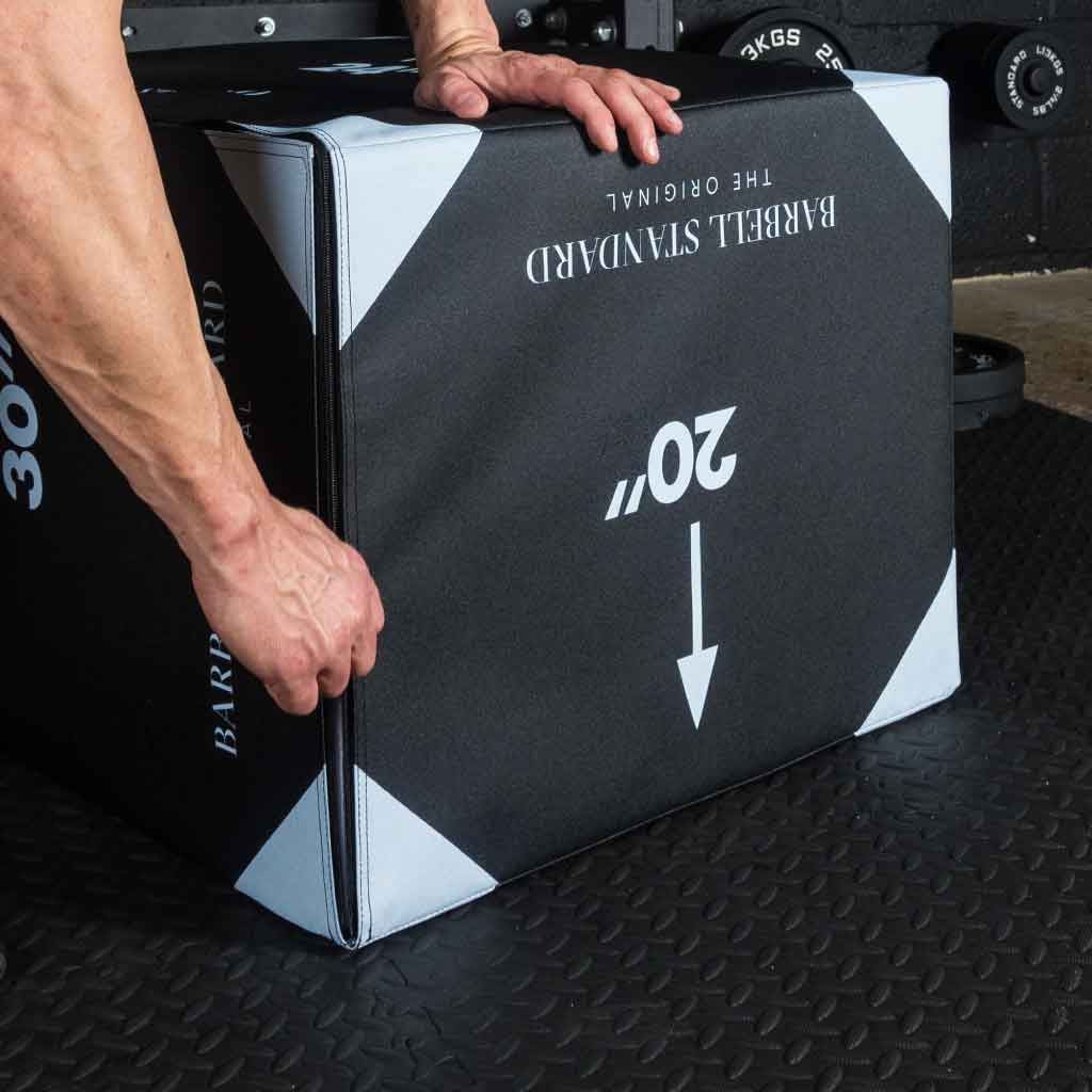 Hand lifting a 20-inch Barbell Standard jump box, black with white arrows.