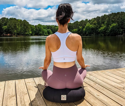 Woman meditating on a wooden dock by a lake; white sports top and mauve leggings.