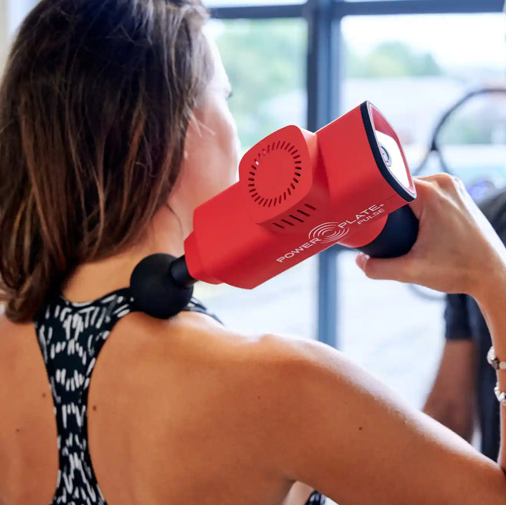 Woman using a red Power Plate massage gun on their shoulder