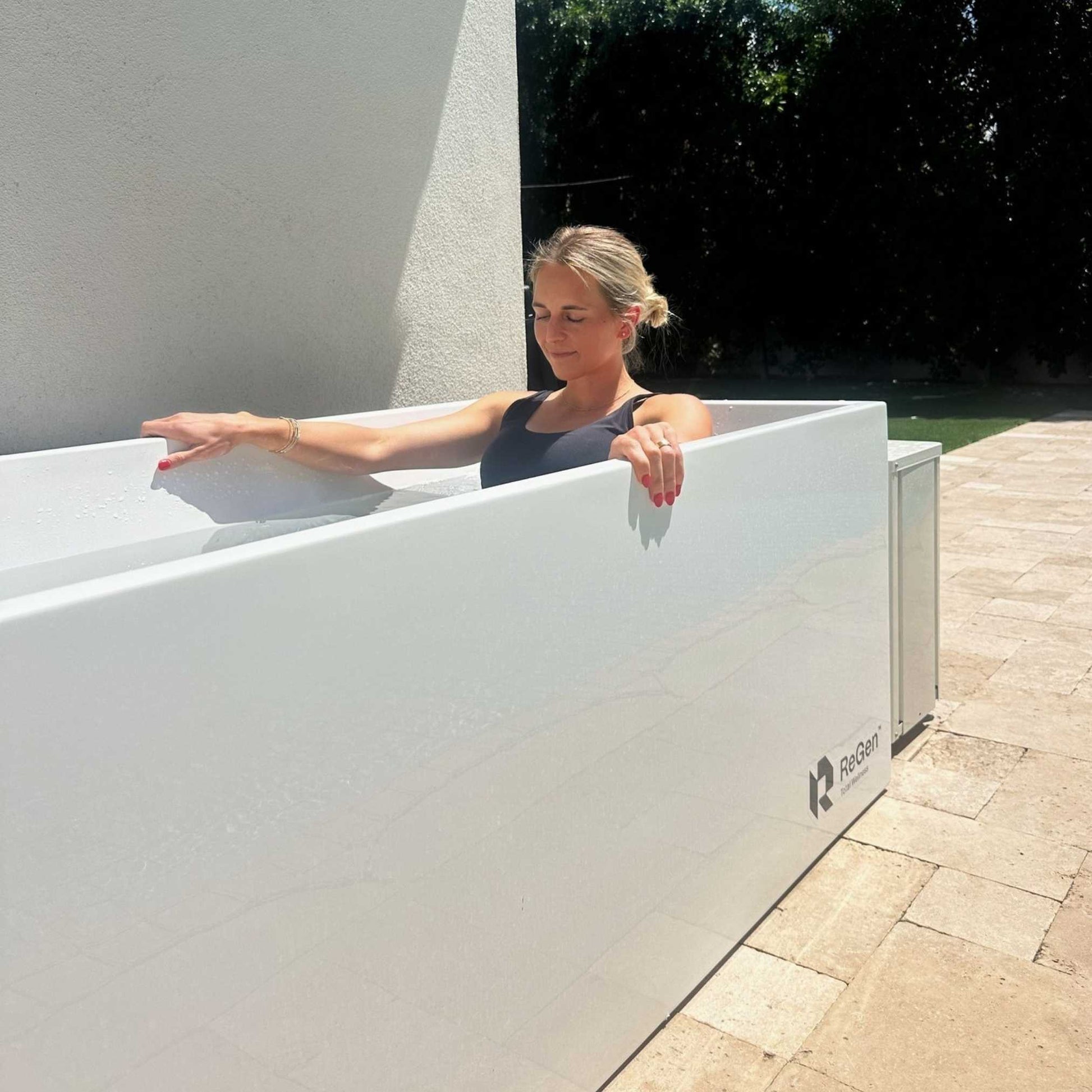 Woman relaxing in a white ReGen ice bath outdoors, enjoying the sun and water for recovery.