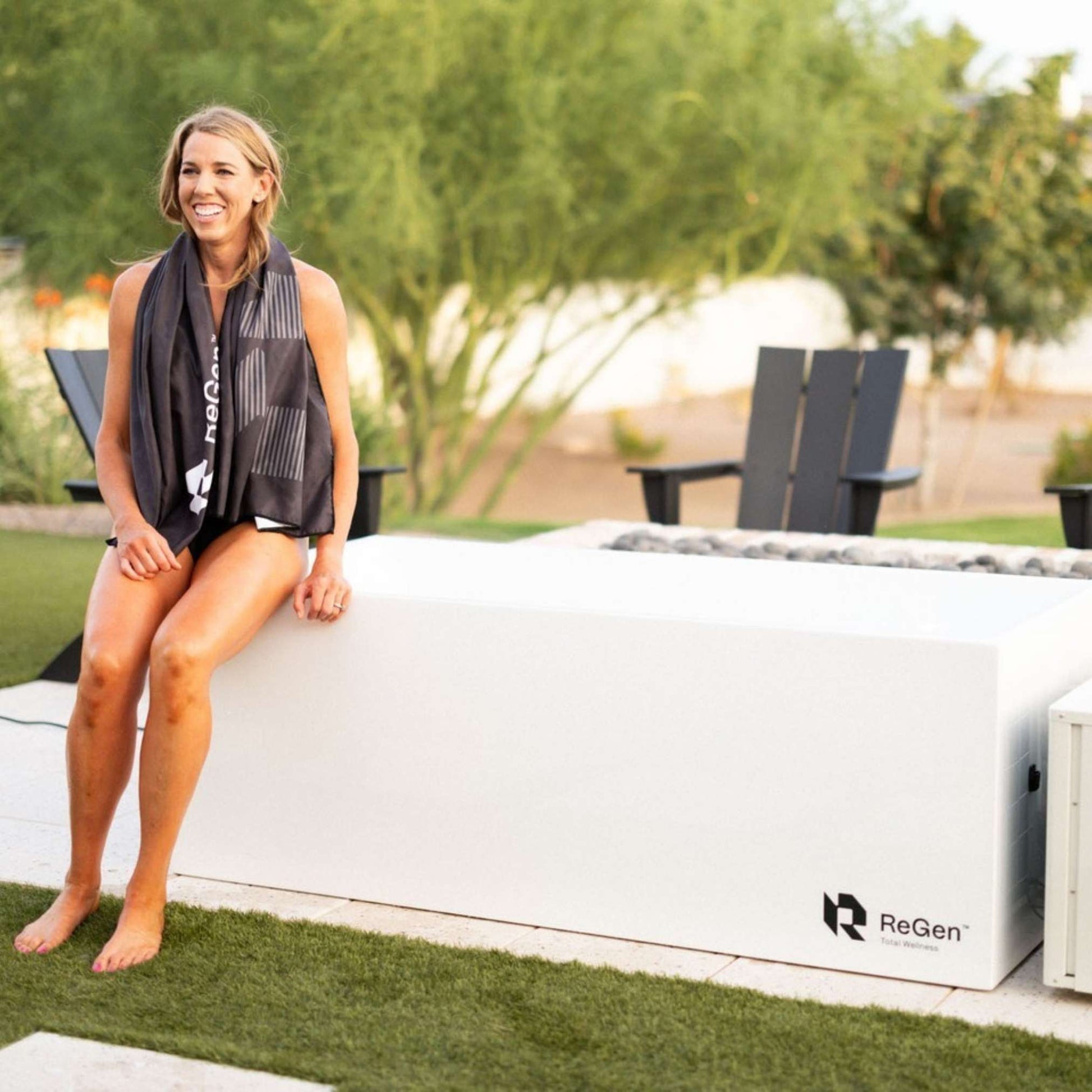 Smiling woman sits on a white ReGen total wellness unit in a backyard setting, wearing a black scarf.