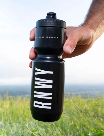Man's hand holding a black water bottle with white "RNWY" text and "Fuel Your Journey" tagline, outdoors with green hills in the background.