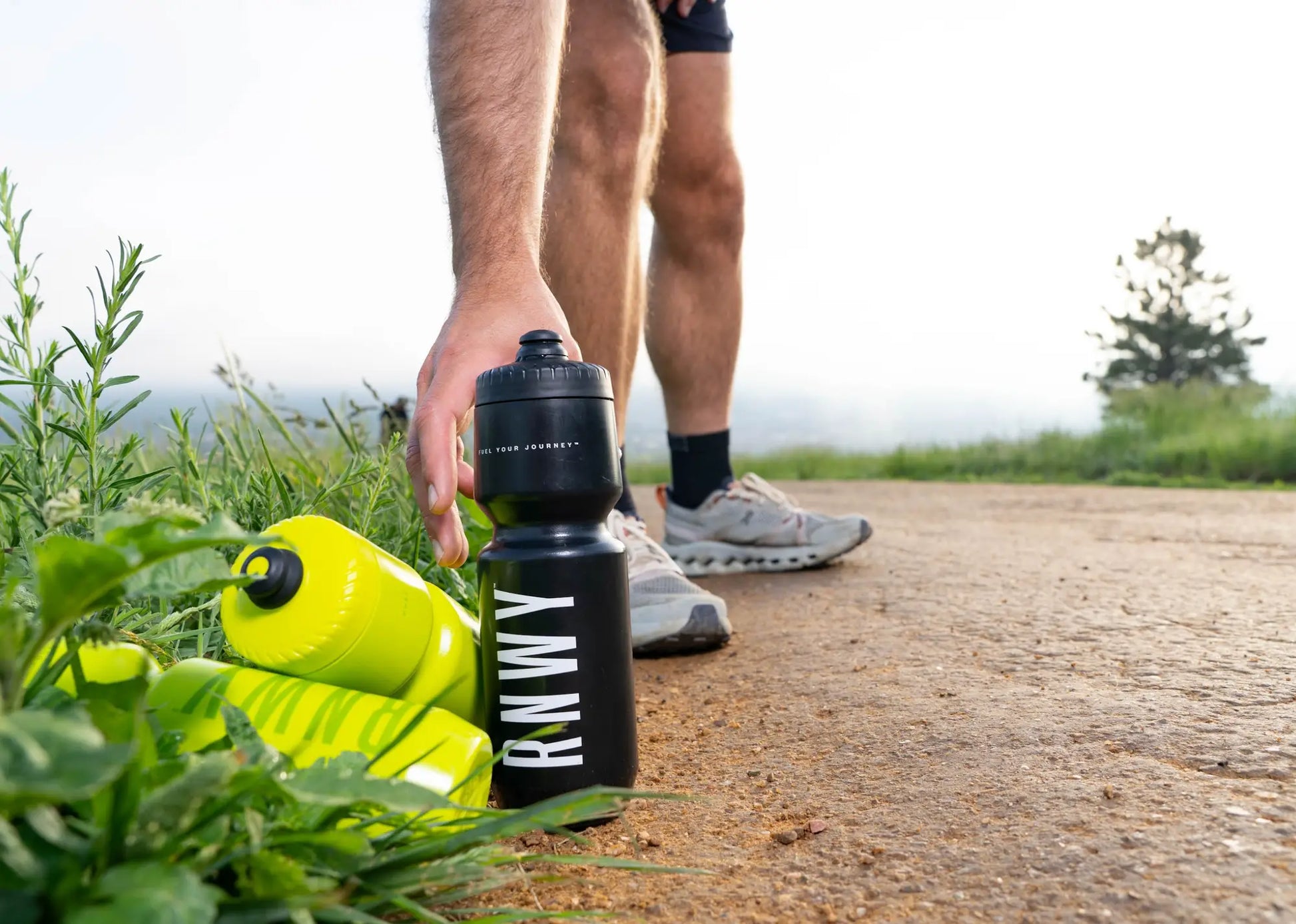 Man picking up a black water bottle labeled "RUNWM" during an outdoor run on a dirt path, with a bright yellow water bottle nearby and green landscape in the background.