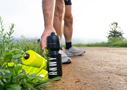 Man picking up a black water bottle labeled "RUNWM" during an outdoor run on a dirt path, with a bright yellow water bottle nearby and green landscape in the background.