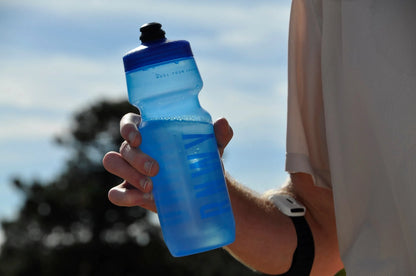 Person holding a blue sports water bottle with blue cap and ribbed grip design outdoors in sunlight.