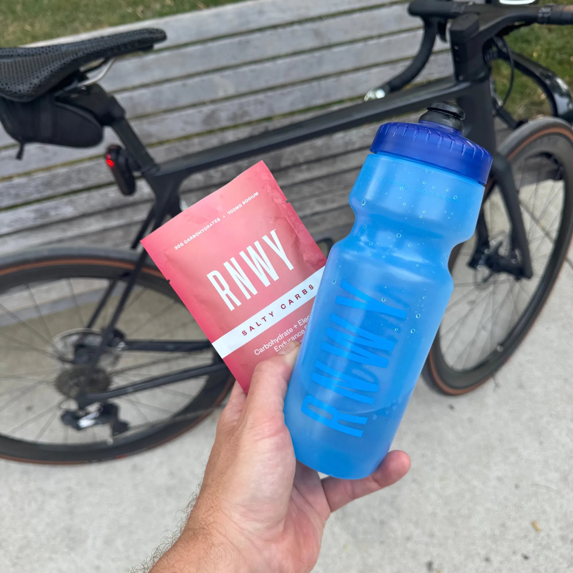 Hand holding a blue 24oz sports water bottle and pink carbohydrate supplement packet in front of a black road bike on a wooden deck.