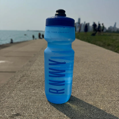 Bright blue sports water bottle with dark blue cap and text graphic on concrete path near a waterfront.