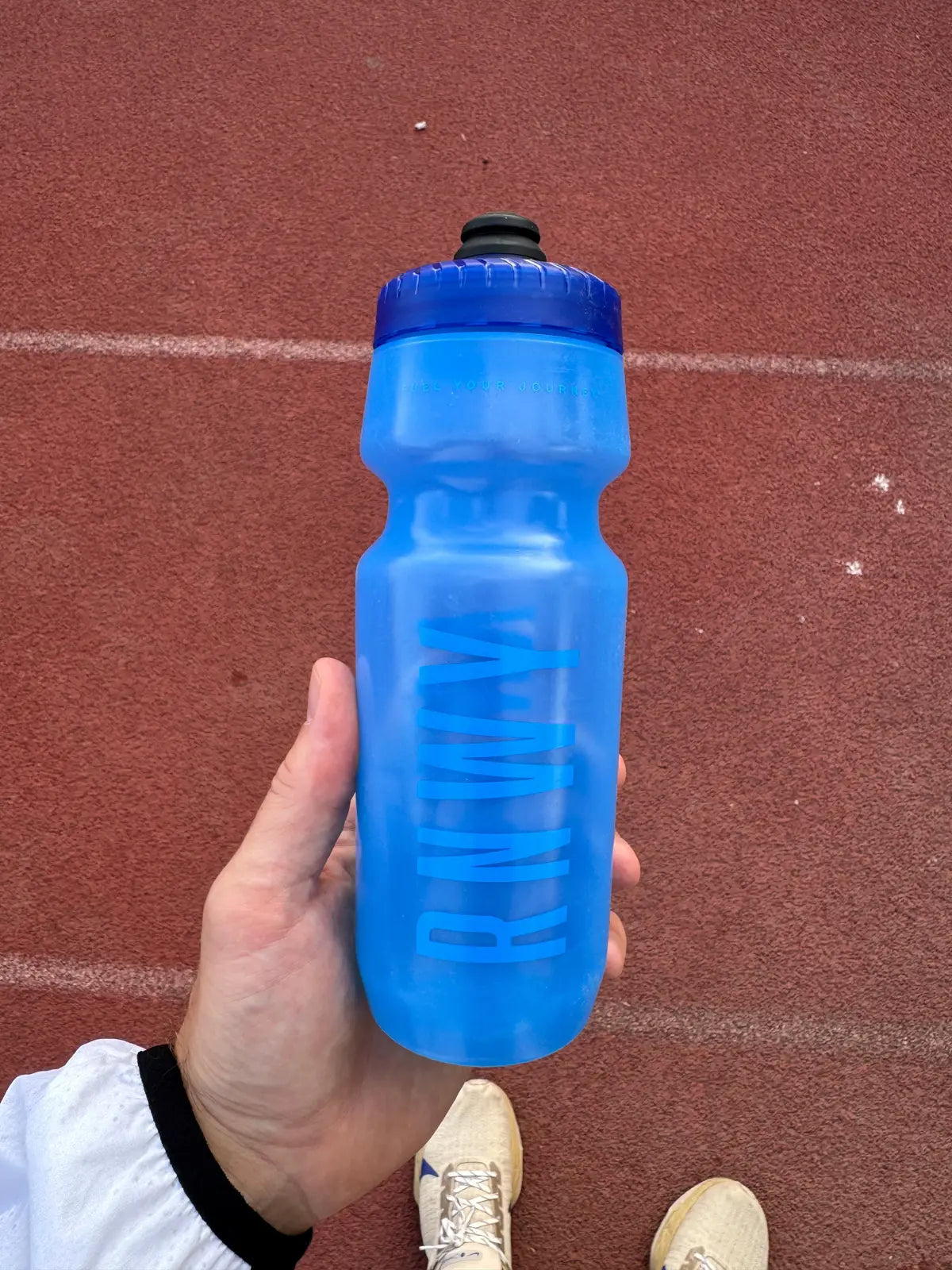 Person holding a blue 24-ounce sports water bottle with a screw-top cap and textured grip, photographed outdoors on a red running track.