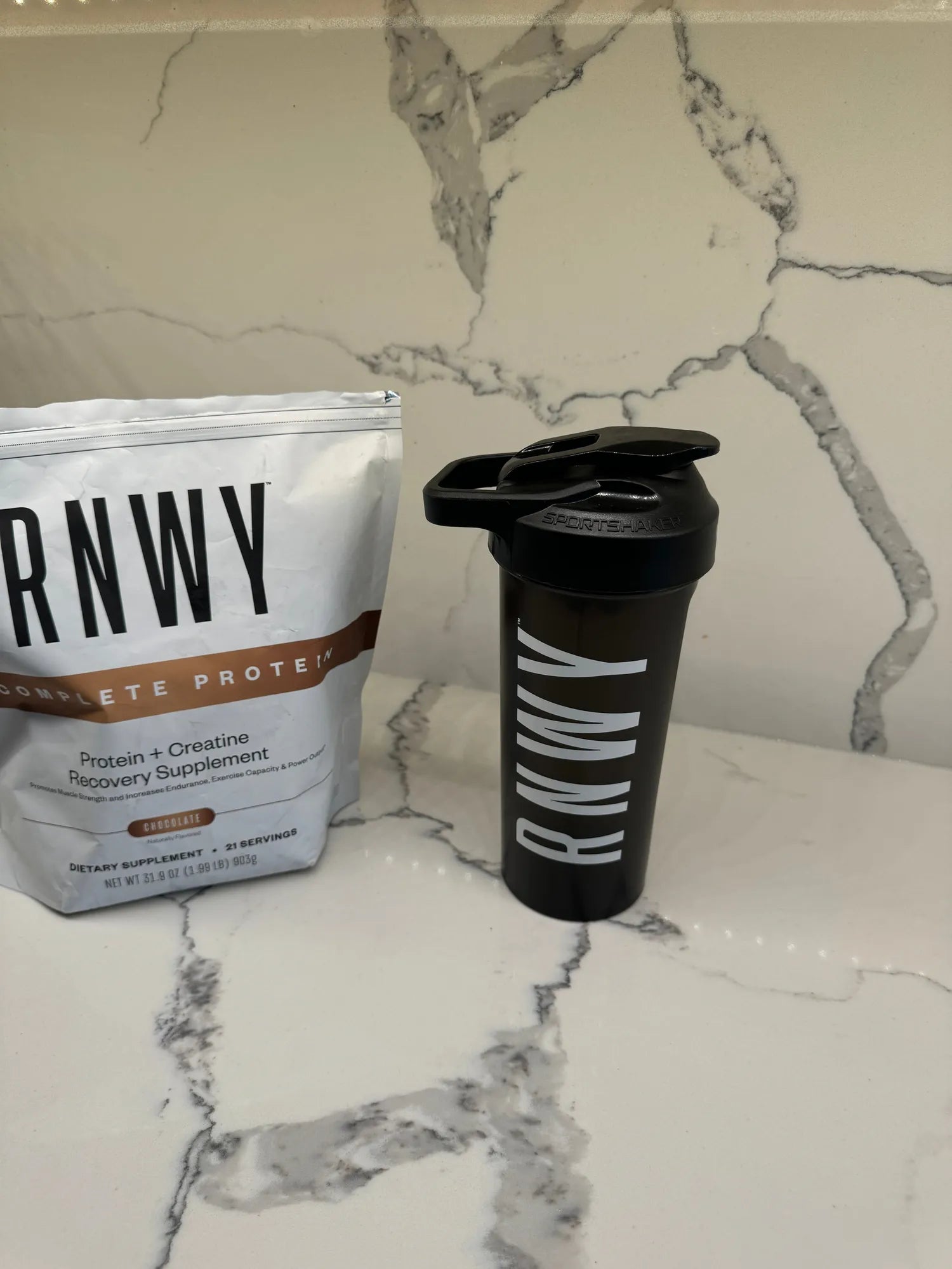 Black protein shaker bottle with white stripe design next to a white resealable pouch of protein powder supplement on a marble surface.