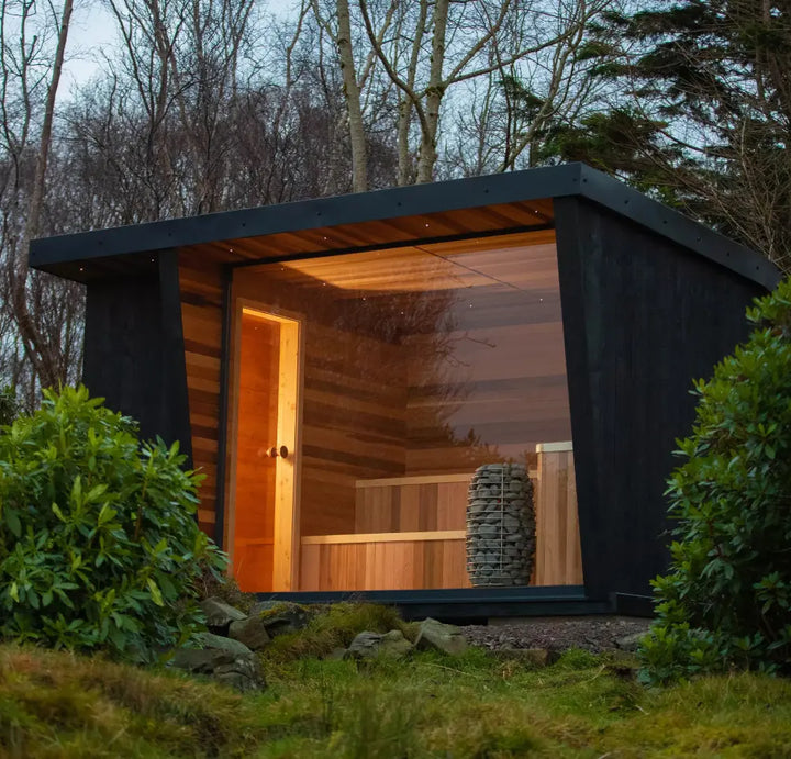 Modern traditional sauna with black exterior and wooden interior, surrounded by greenery.