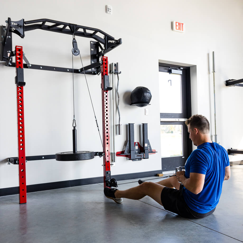 Man in blue shirt sits on gym floor beside red PRX cable machine and attachments.