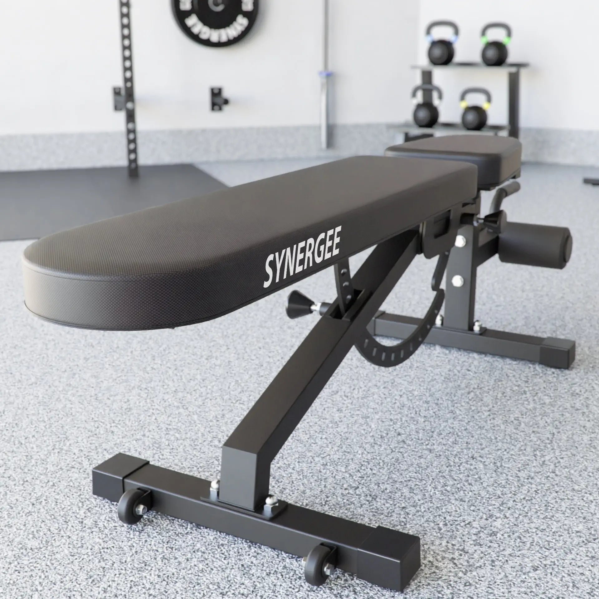 Black adjustable weight bench with "Synergee" branding on the padded seat, featuring an incline-decline mechanism and foot platform. A gym setting with kettlebells on a rack and wall-mounted equipment visible in the background.