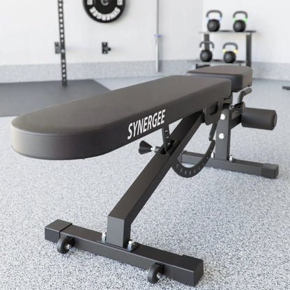 Black adjustable weight bench with "Synergee" branding on the padded seat, featuring an incline-decline mechanism and foot platform. A gym setting with kettlebells on a rack and wall-mounted equipment visible in the background.