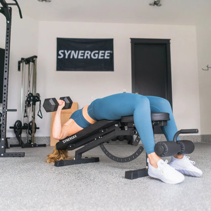 Woman performing dumbbell chest press on a black adjustable incline-decline bench in a gym setting with dumbbells and power rack visible in the background.
