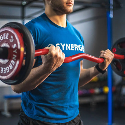 Man performing a bicep curl with a red curved barbell in a gym setting with weight plates visible in the background.