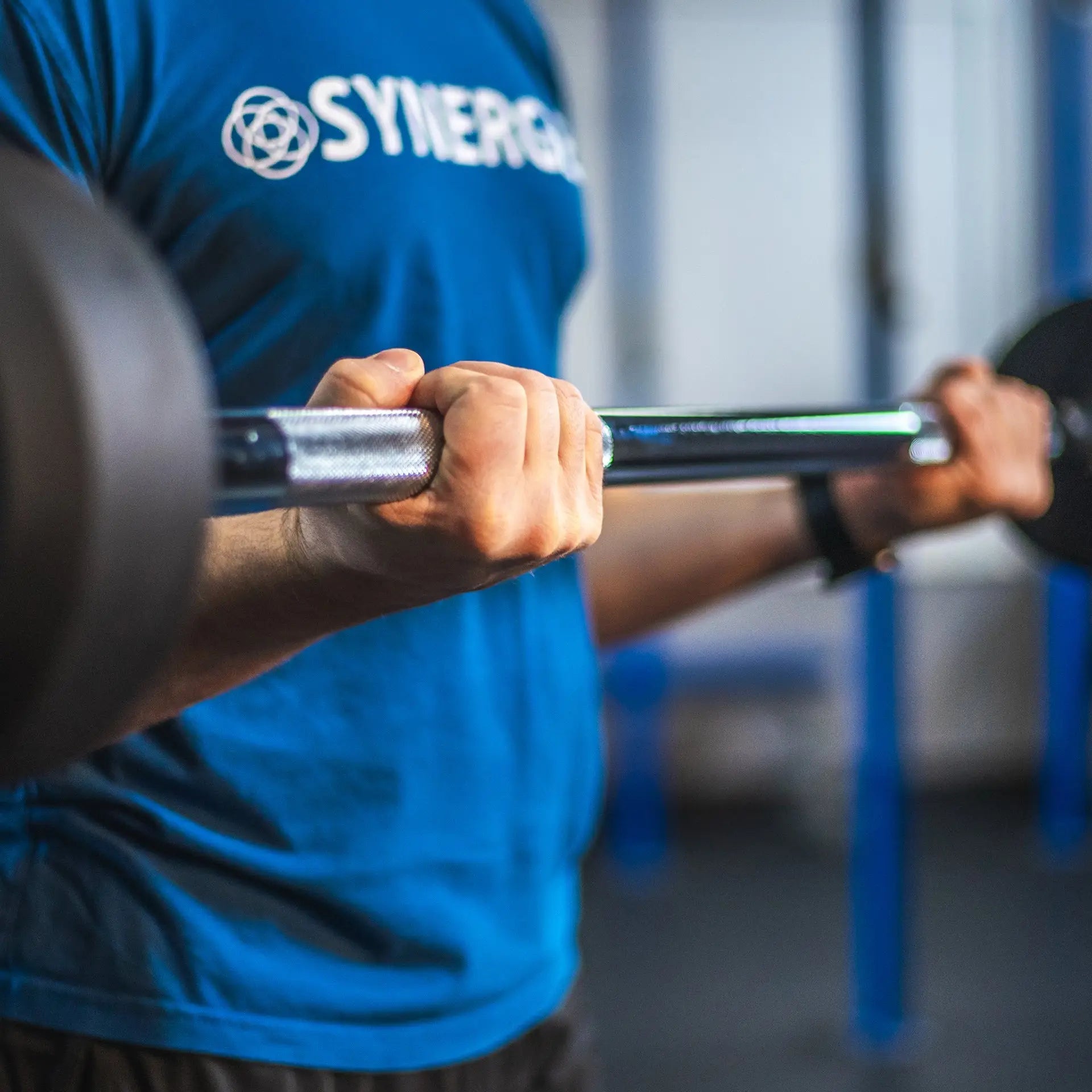 Person gripping a fixed barbell with green and black knurled sleeves in a gym setting, wearing a blue Synergee branded shirt.