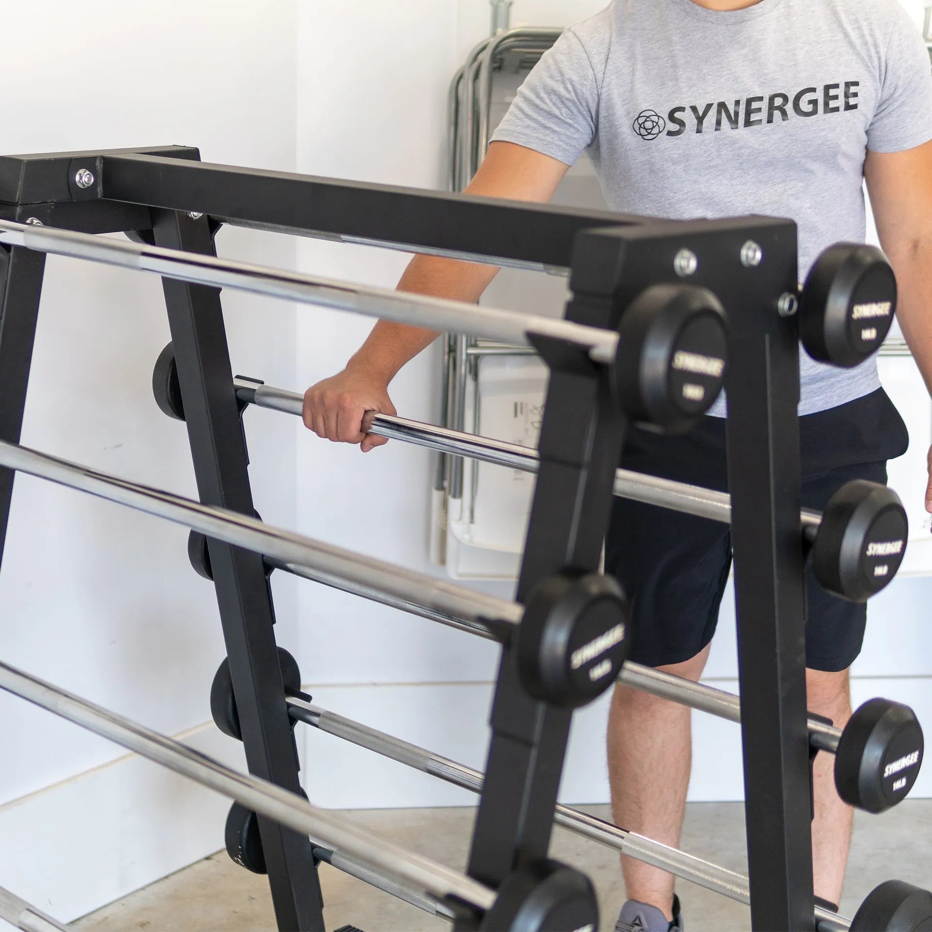 Man holding a chrome barbell in front of a black fixed barbell storage rack with multiple pairs of black dumbbells mounted on horizontal bars.