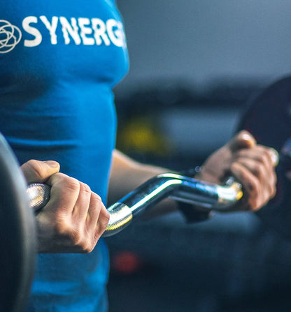 Person in blue Synerg shirt holding a fixed curved barbell with black bar and yellow grip accents.