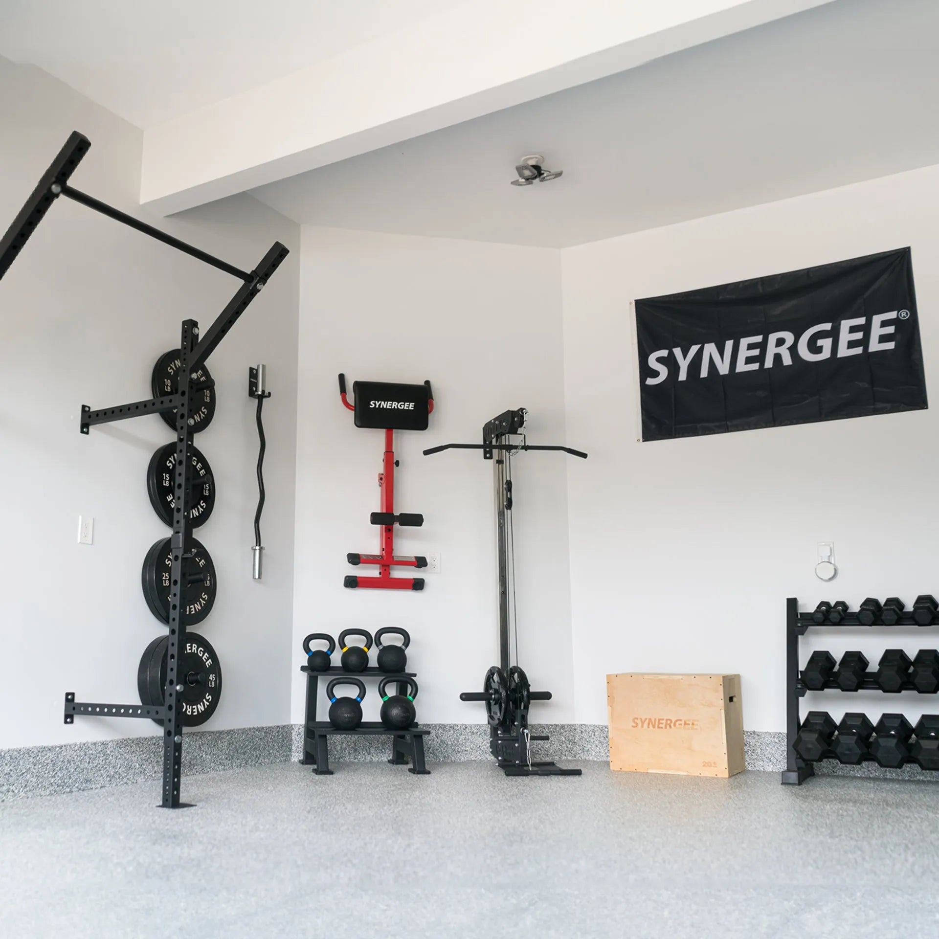 Black Synergee flag banner displayed on white gym wall above assorted fitness equipment including wall-mounted barbell rack, kettlebells, red pull-up bar, and dumbbell rack.
