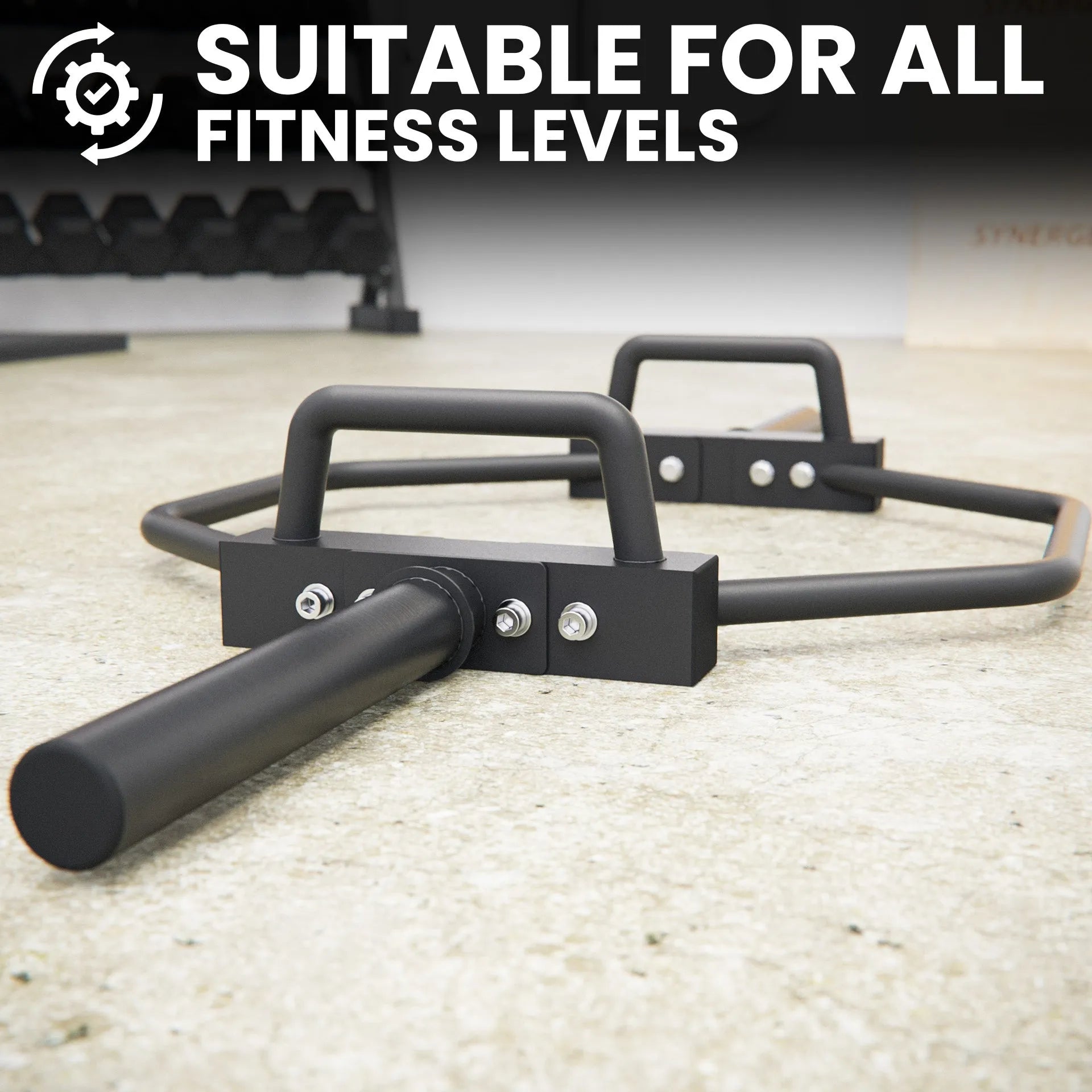 Black hex trap bar with curved handles and metal attachment points, displayed on a light concrete gym floor with dumbbells in the background.